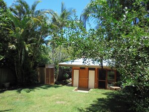 A small wooden building with a door and windows, surrounded by lush greenery and palm trees.