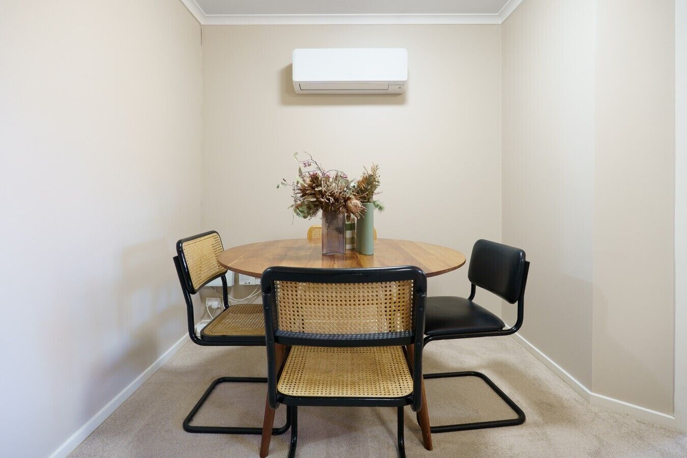 A room with a round wooden table, four chairs, a vase with dried flowers, and an air conditioning unit on the wall.