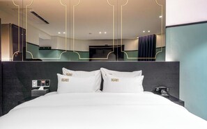 Habitación de hotel con una cama grande, dos almohadas bordadas con la palabra 'AGAIN' y una mesita de noche con un teléfono, además de un espejo que refleja la habitación.