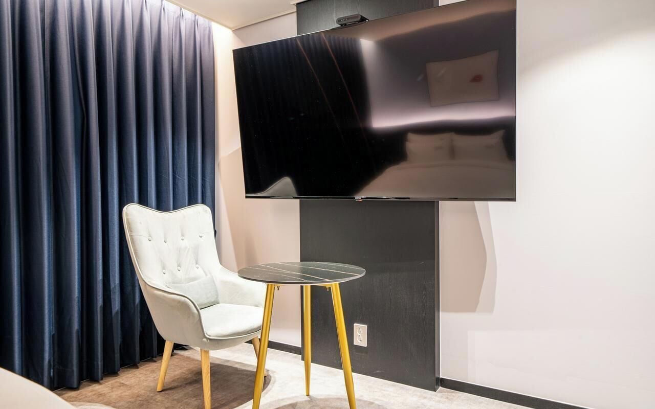 A room with a flat-screen TV mounted on the wall, a white tufted chair, and a small round table with yellow legs.