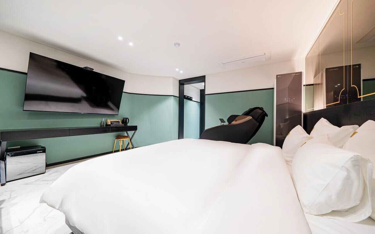 A modern hotel room with a large bed, a flat-screen TV mounted on the wall, a desk with a chair, and a black lounge chair.
