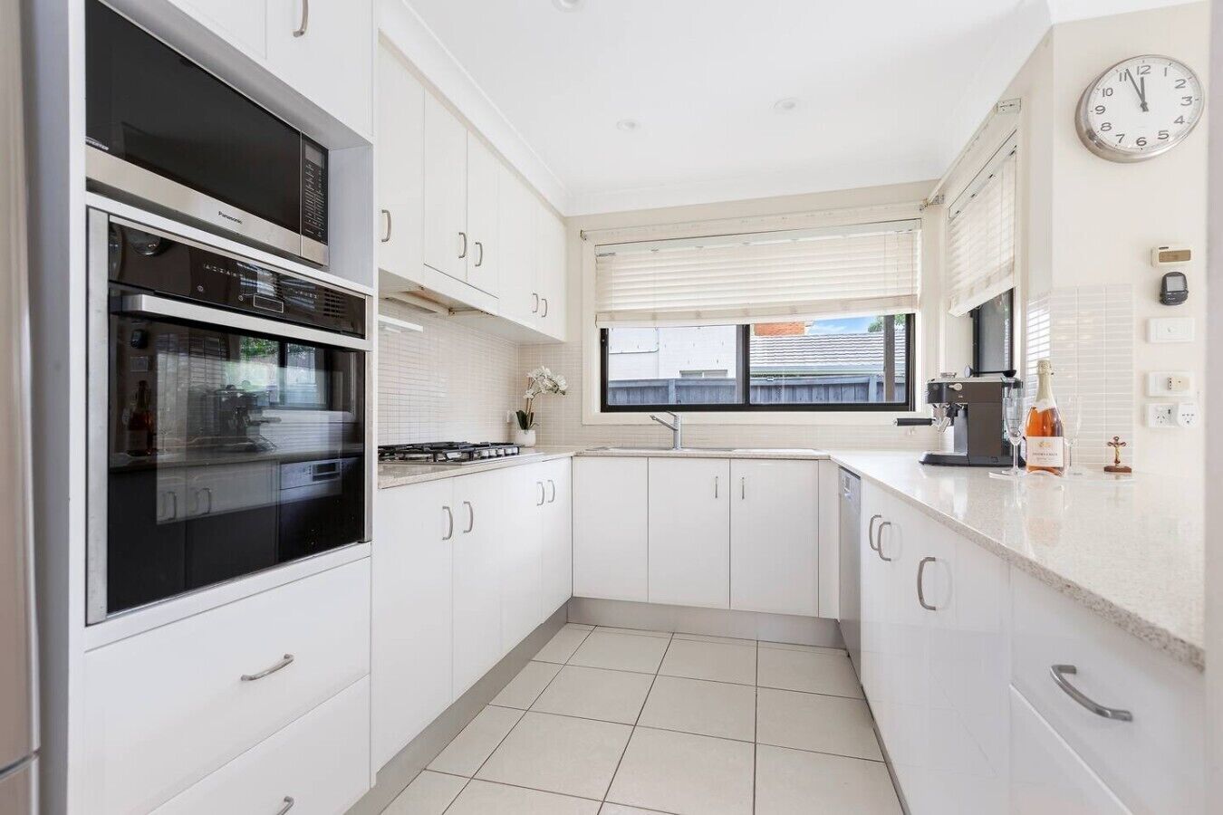 A modern kitchen with white cabinetry, a built-in oven, a window with blinds, and a clock on the wall.