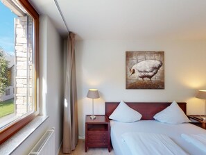 A hotel room with a bed, two bedside tables, a lamp, a painting of a bird, and a window with curtains.