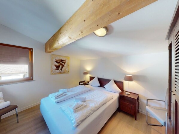 A bedroom with a bed, two bedside tables, a nightstand, a window with blinds, and a wooden ceiling beam.