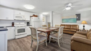 A modern kitchen with white appliances, a dining area with a wooden table and chairs, and a living room with a brown leather sofa. - Beach Please! · Ocean View 2BR · Cherry Grove (North Myrtle Beach)