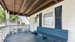A covered porch with a wooden bench and white chairs, a wooden ceiling, and white walls with shutters. - Beach Please! · Ocean View 2BR · Cherry Grove (North Myrtle Beach)