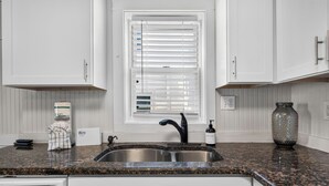 A kitchen with white cabinets, a granite countertop, a double sink, a window with blinds, and a vase on the counter. - Beach Please! · Ocean View 2BR · Cherry Grove (North Myrtle Beach)