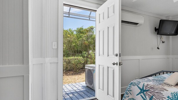 An open door leading to a balcony with an air conditioning unit, a bed with a starfish-patterned bedspread, and a mounted television. - Beach Please! · Ocean View 2BR · Cherry Grove (North Myrtle Beach)