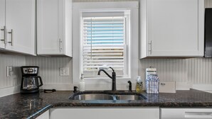 A kitchen with white cabinets, a double sink, a coffee maker, and a window with blinds.