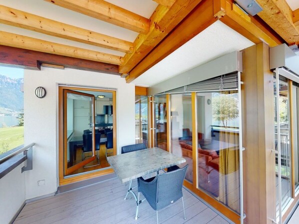 A modern outdoor dining area with a table and chairs, wooden beams overhead, and a glass door leading to an interior space.