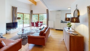 Ein modernes Wohnzimmer mit einer Couch, einem Fernseher, einem Esstisch mit Stühlen, einer Küchenzeile und großen Fenstern.