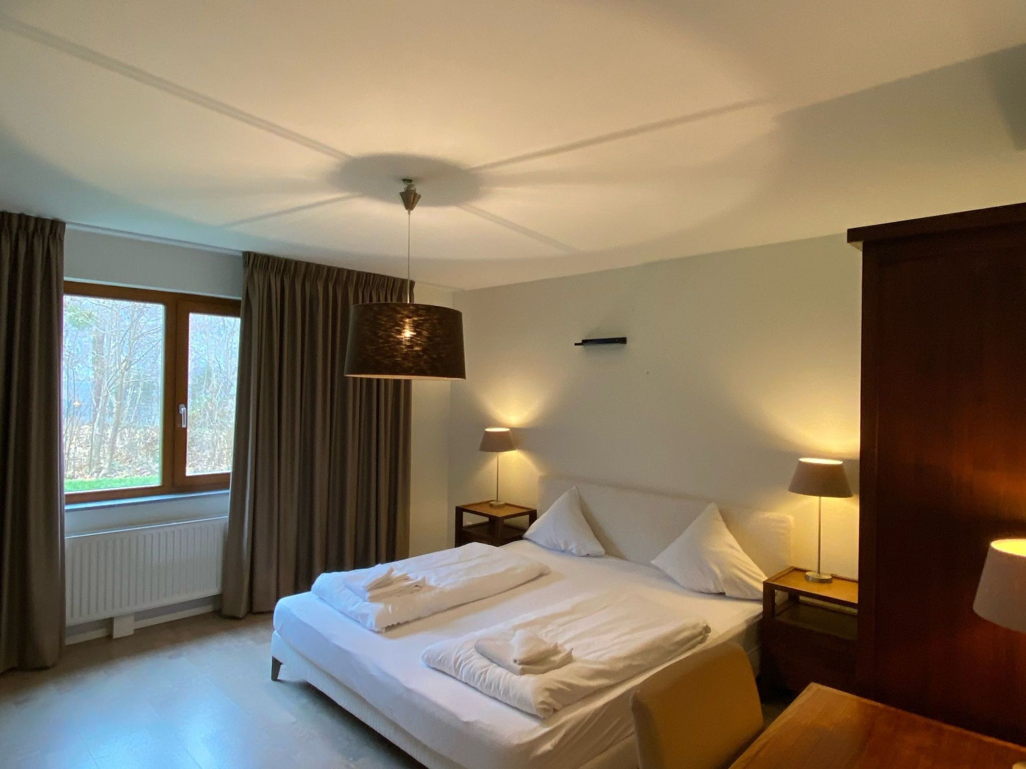 A hotel room with a bed, two bedside lamps, a window with curtains, and a radiator.