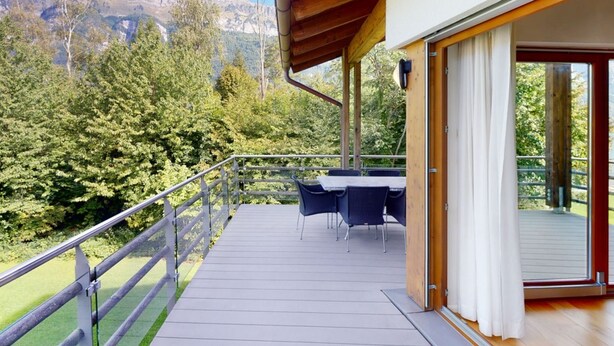 Een houten terras met buitenmeubilair, een glazen deur naar een overdekt gedeelte en uitzicht op een berg.