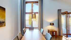 Een moderne eetruimte met een lange houten tafel, stoelen, een hanglamp en uitzicht door grote ramen.