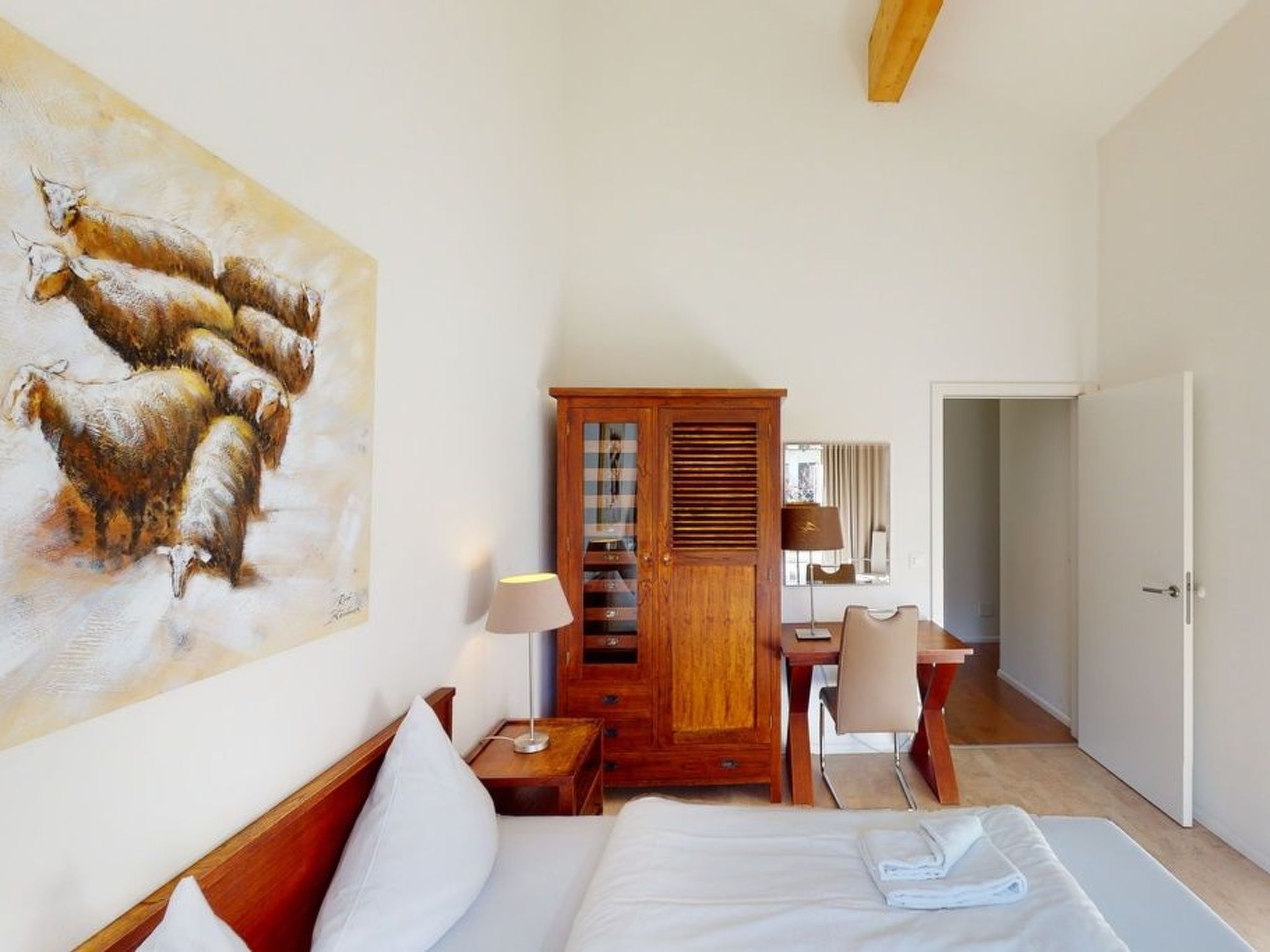 A hotel room with a bed, a wooden wardrobe, a desk with a chair, and a painting of sheep on the wall.