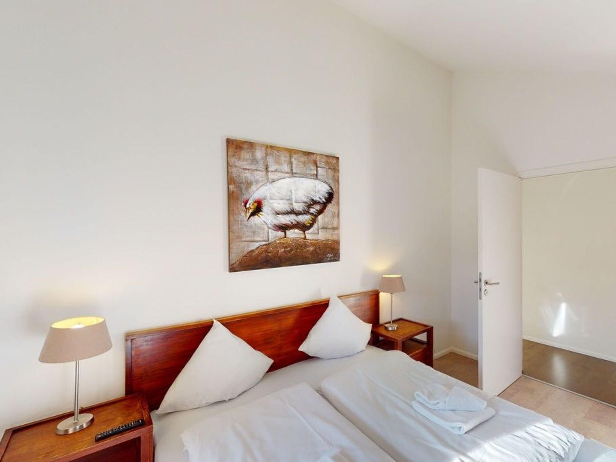 Habitación de hotel con cama, dos mesitas de noche, una lámpara, un cuadro de un pájaro y una puerta.