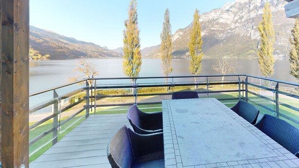 Een terras met een tafel en stoelen, met uitzicht op een meer en bergen.