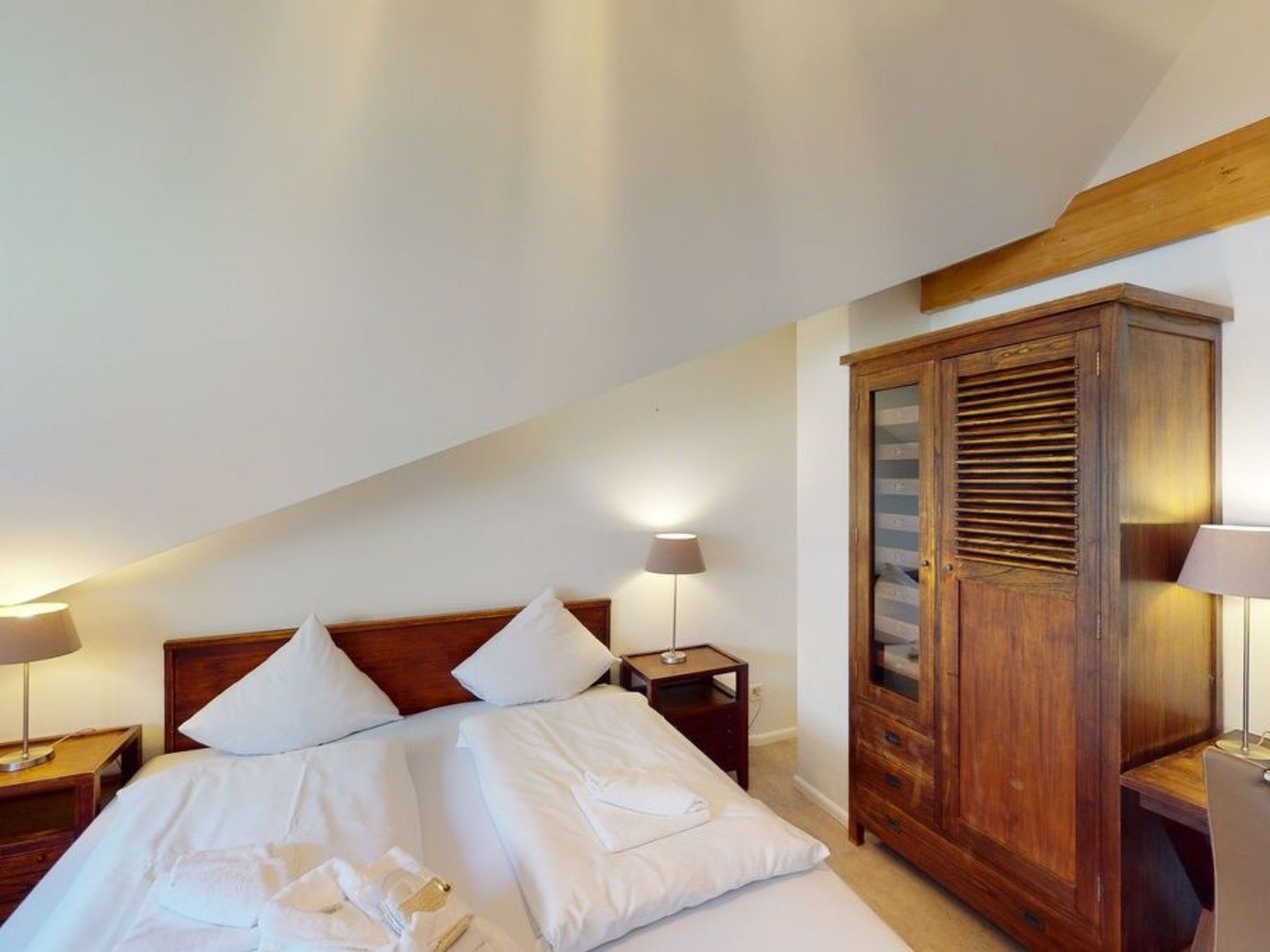 A bedroom with a bed, two bedside tables, a wooden wardrobe, and a slanted ceiling.