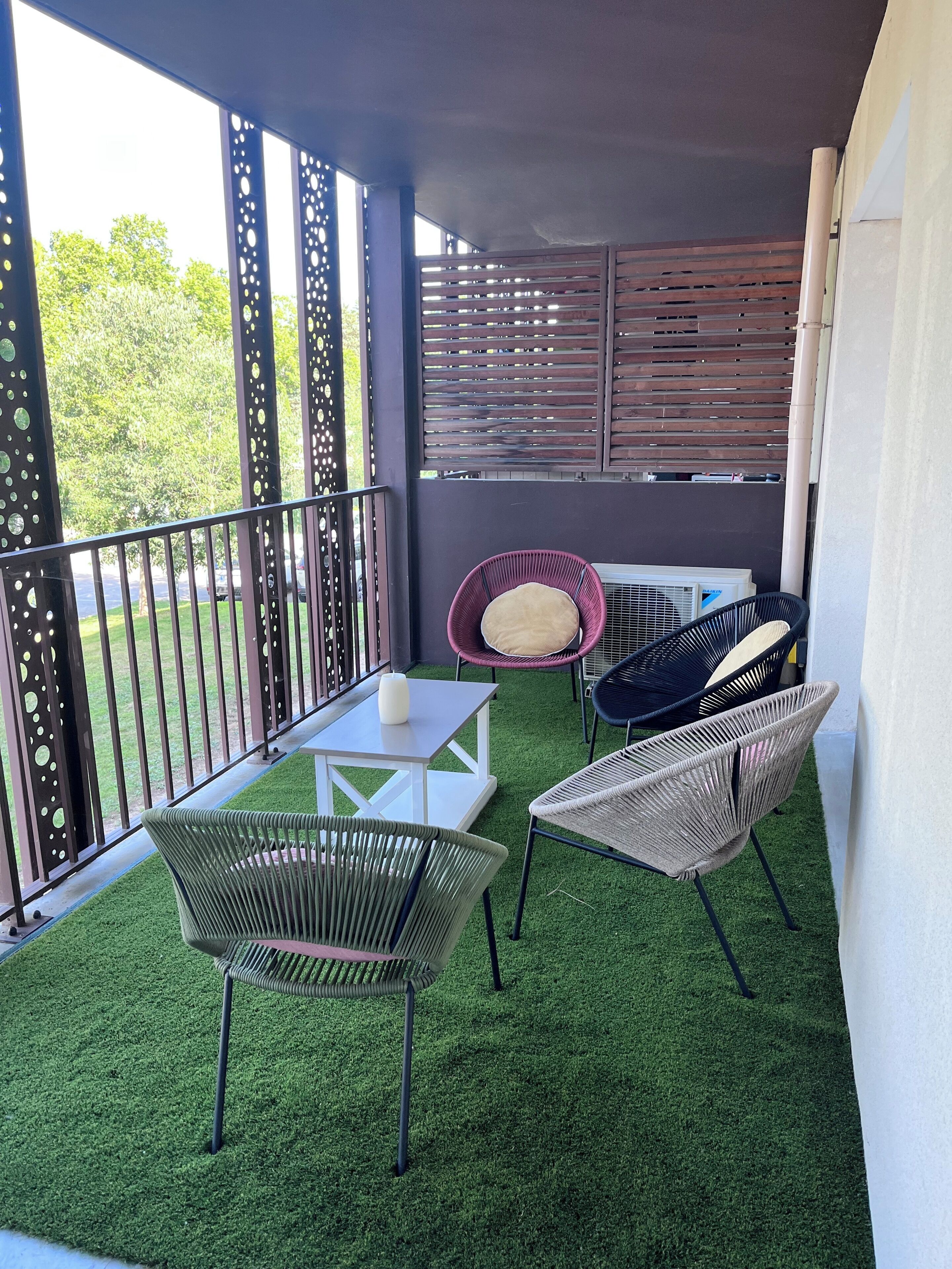 Un balcón con césped artificial, sillas de mimbre, una mesa blanca y una planta en maceta.