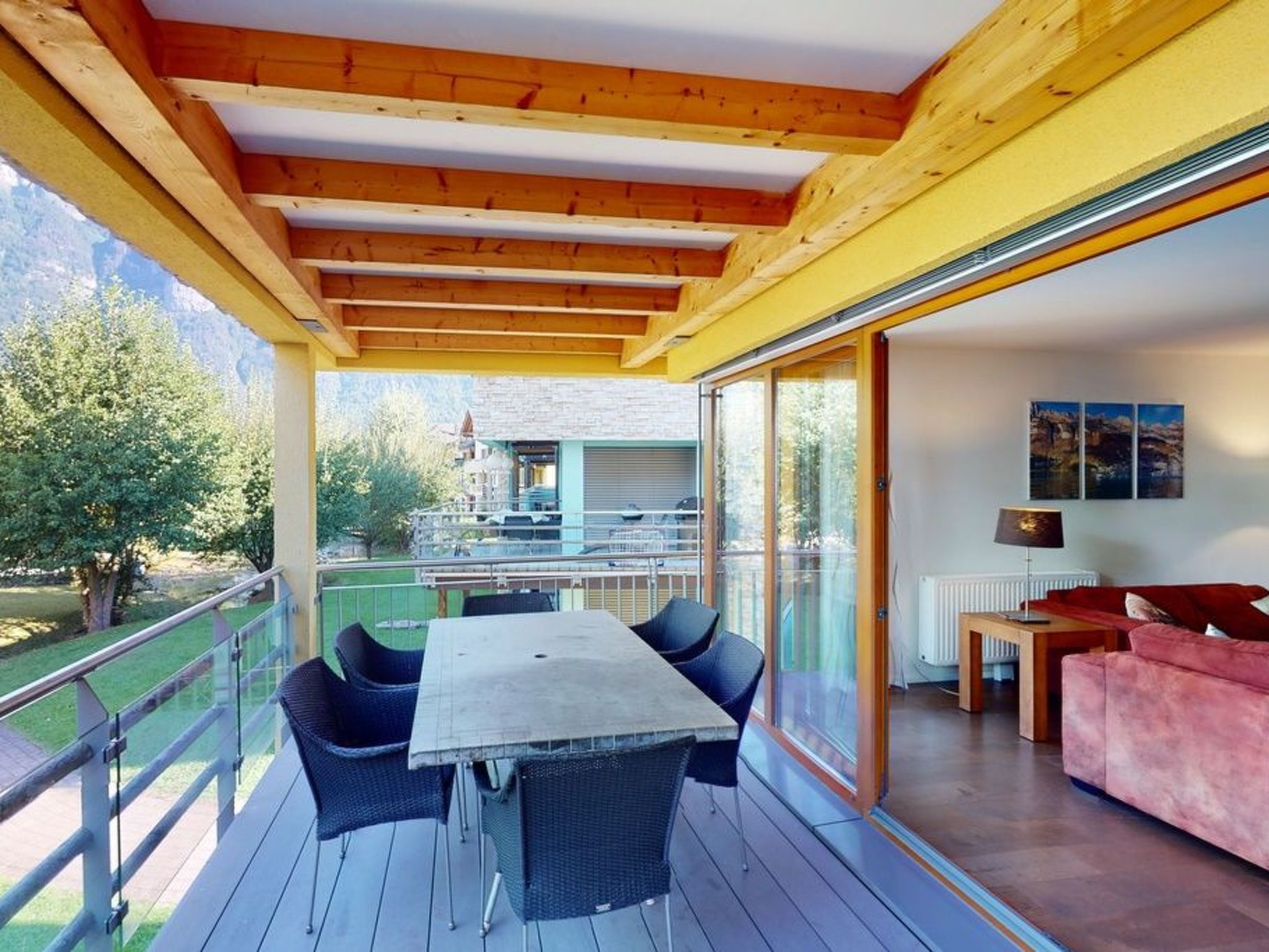Un espacio exterior cubierto con mesa y sillas, un sofá y vistas a un edificio y árboles.