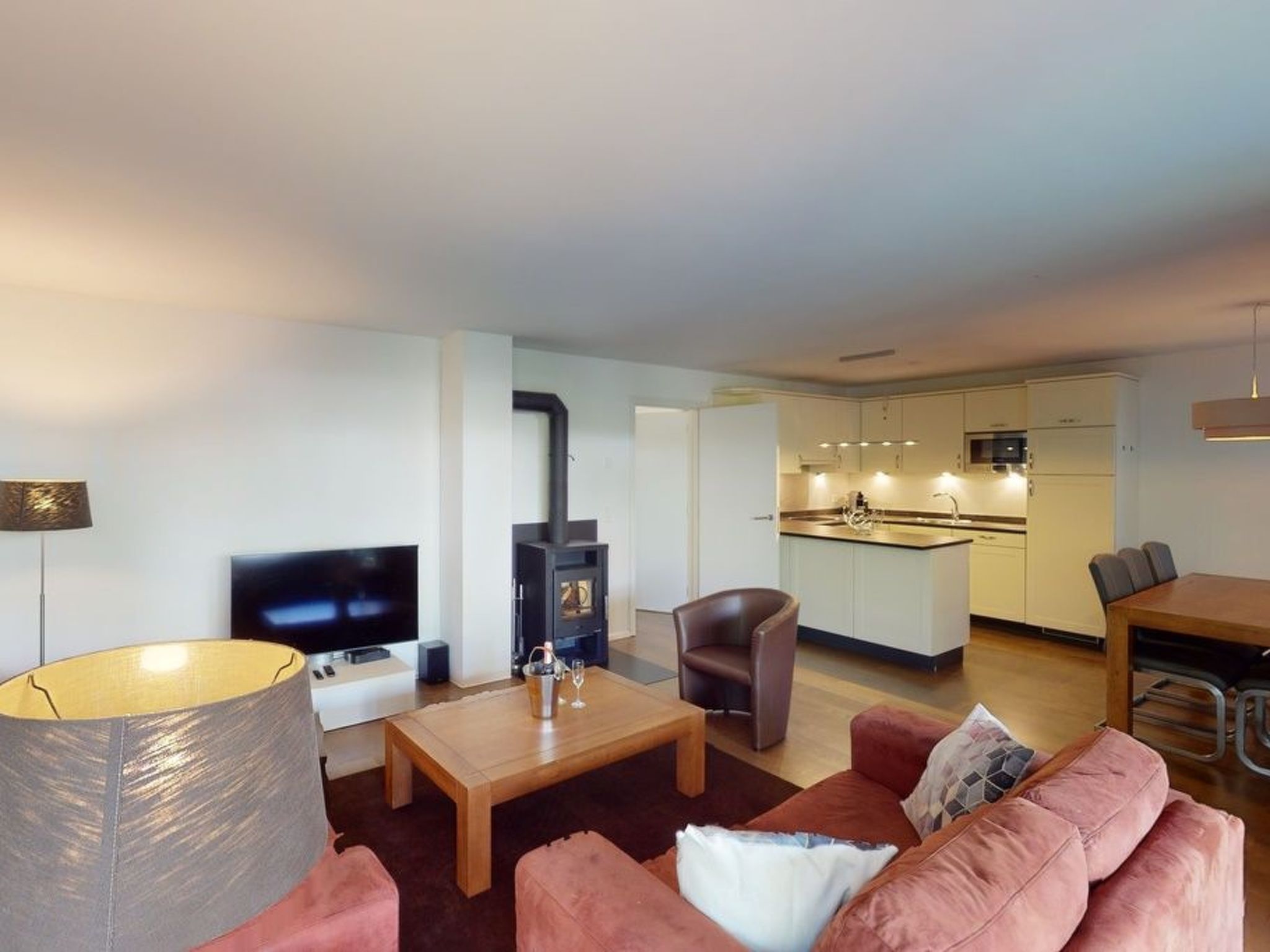 A modern living room with a flat-screen TV, a fireplace, a dining area with a table and chairs, and a kitchen with white cabinets and a countertop.