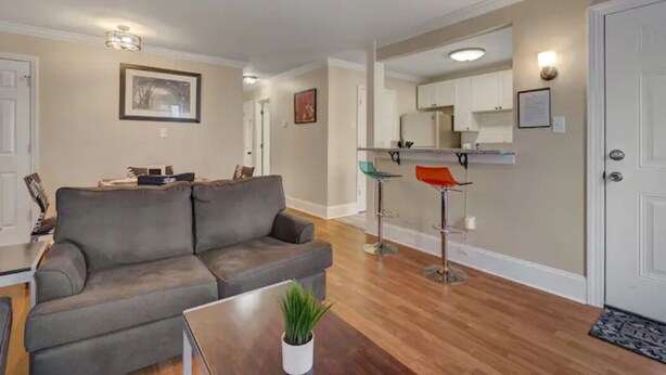 Una sala de estar con un sofá gris, una mesa de centro y una planta. Se observa una zona de cocina con armarios blancos y una encimera a través de un vano abierto.