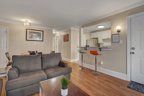 A living room with a gray sofa, a coffee table, and a plant. There is a kitchen area with white cabinets and a countertop visible through an open doorway. - 2-bedroom apartment in tranquil Durham with cool AC breeze (Durham)