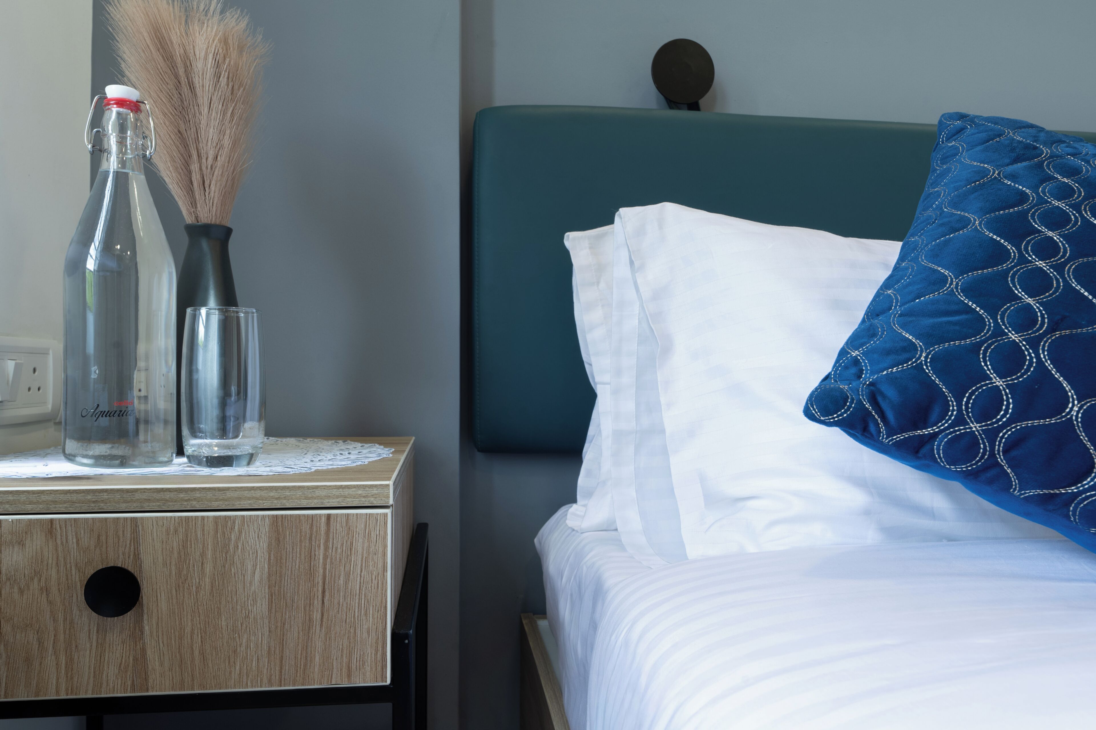 Una cama bien hecha con sábanas blancas y una almohada de estampado azul, una mesita de noche con un frasco transparente y un vaso, y una mesita de noche fijada a la pared.