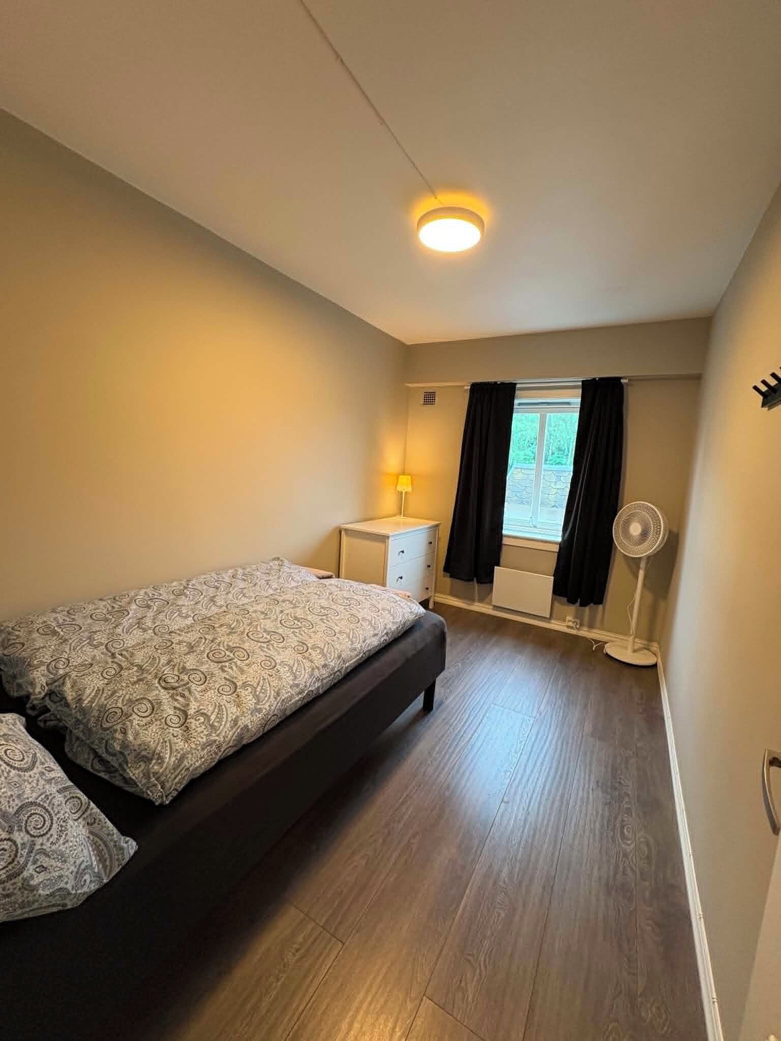 A bedroom with a bed, bedside table, lamp, window with curtains, and a standing fan.