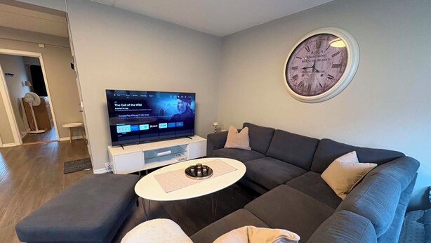 A modern living room with a sectional sofa, a flat-screen TV, a round wall clock, and a small coffee table. - Notodden Sentrum Apartment NO 14 (Telemark)