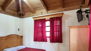 Habitació amb dos llits de fusta, un escriptori de fusta, una finestra amb cortines vermelles i un sostre de fusta amb bigues al descobert.