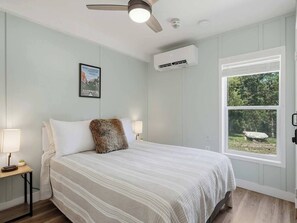 A bedroom with a bed, a ceiling fan, an air conditioning unit, a window with a view of greenery, and a framed picture on the wall. - Private Creekside Cabin • Hot Tub • Sauna • Views (North Fork)