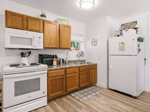 A kitchen with wooden cabinets, a white stove, a microwave, a refrigerator, and a window with blinds.