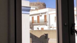 A building with a stone structure on top, a balcony with a metal railing, and a window with a white frame. - The Courtyard by the Seaside (Calasetta)