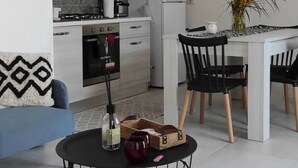 A modern kitchen with a gas stove, oven, and white refrigerator. There is a dining table with black chairs and a vase of yellow flowers. A blue sofa and a small round table with a bottle and two mugs are also visible. - The Courtyard by the Seaside (Calasetta)