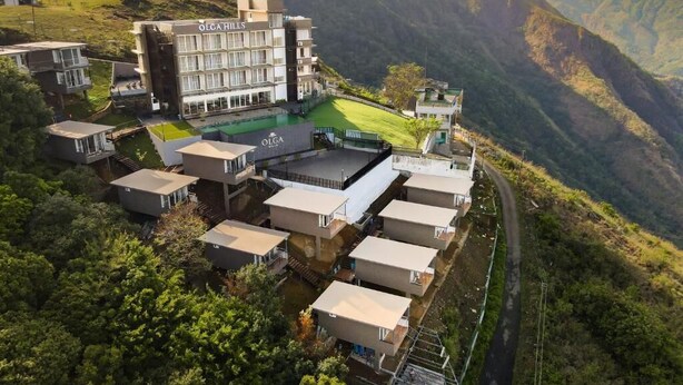 A hotel with a modern design, multiple buildings, and a pool area, surrounded by lush greenery and mountains.