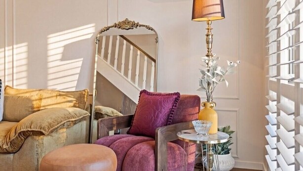 Una sala de estar con sofá, reposapiés, sillón, espejo, lámpara y una mesita con una planta.
