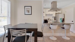 Een eethoek met een tafel en stoelen, een keuken met witte kastjes en een fornuis, en een lijst met een afbeelding aan de muur.