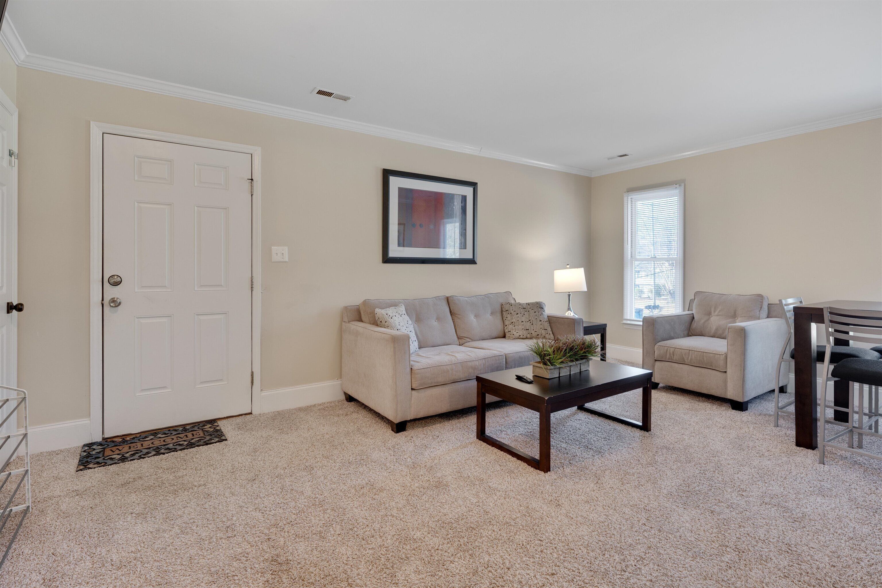 Un salon avec un canapé beige, une table basse et un fauteuil. On y trouve une porte, une fenêtre avec des stores et un tableau encadré au mur.
