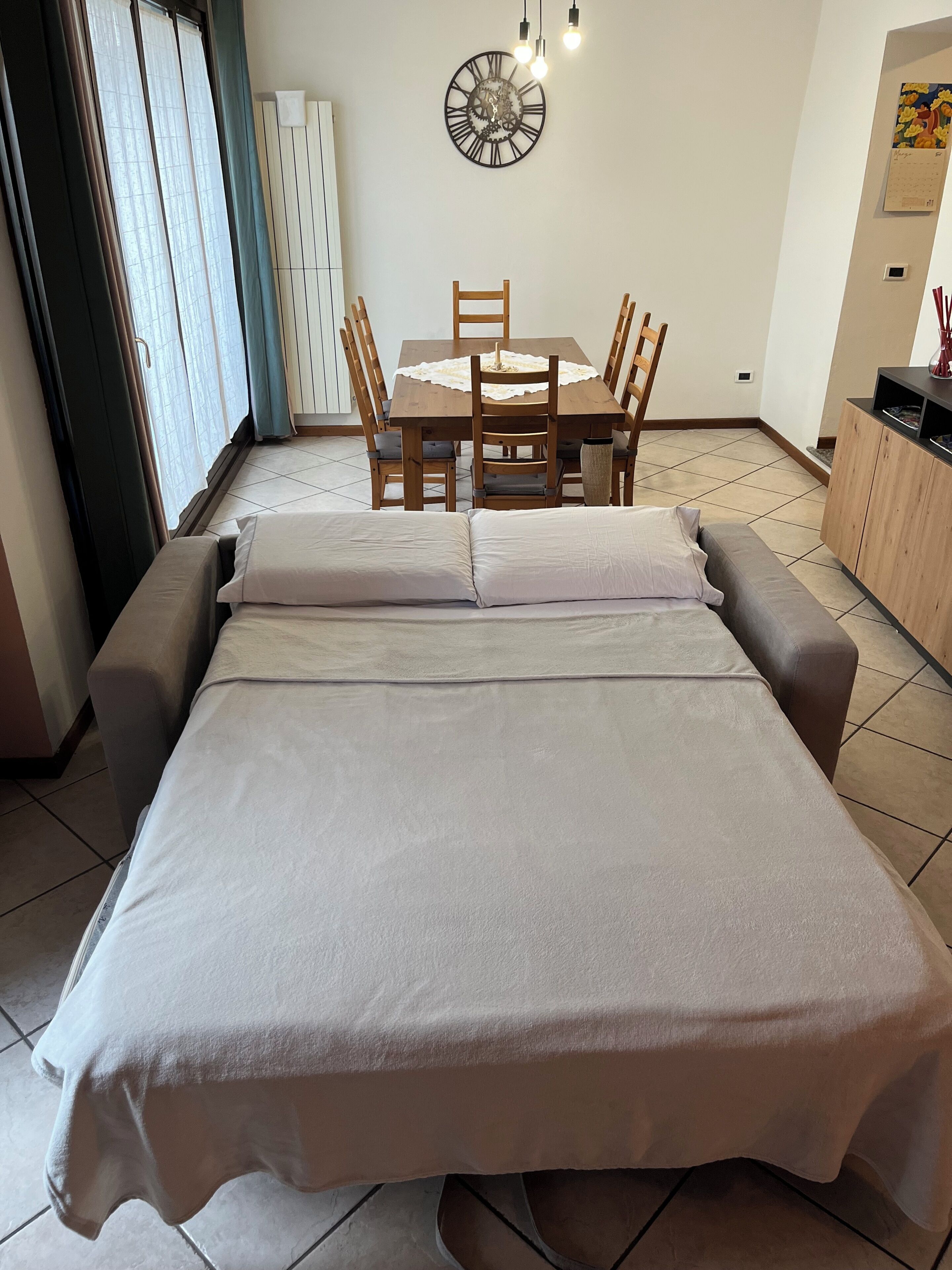 A room with a bed, a dining table with chairs, a wall clock, and a cabinet.
