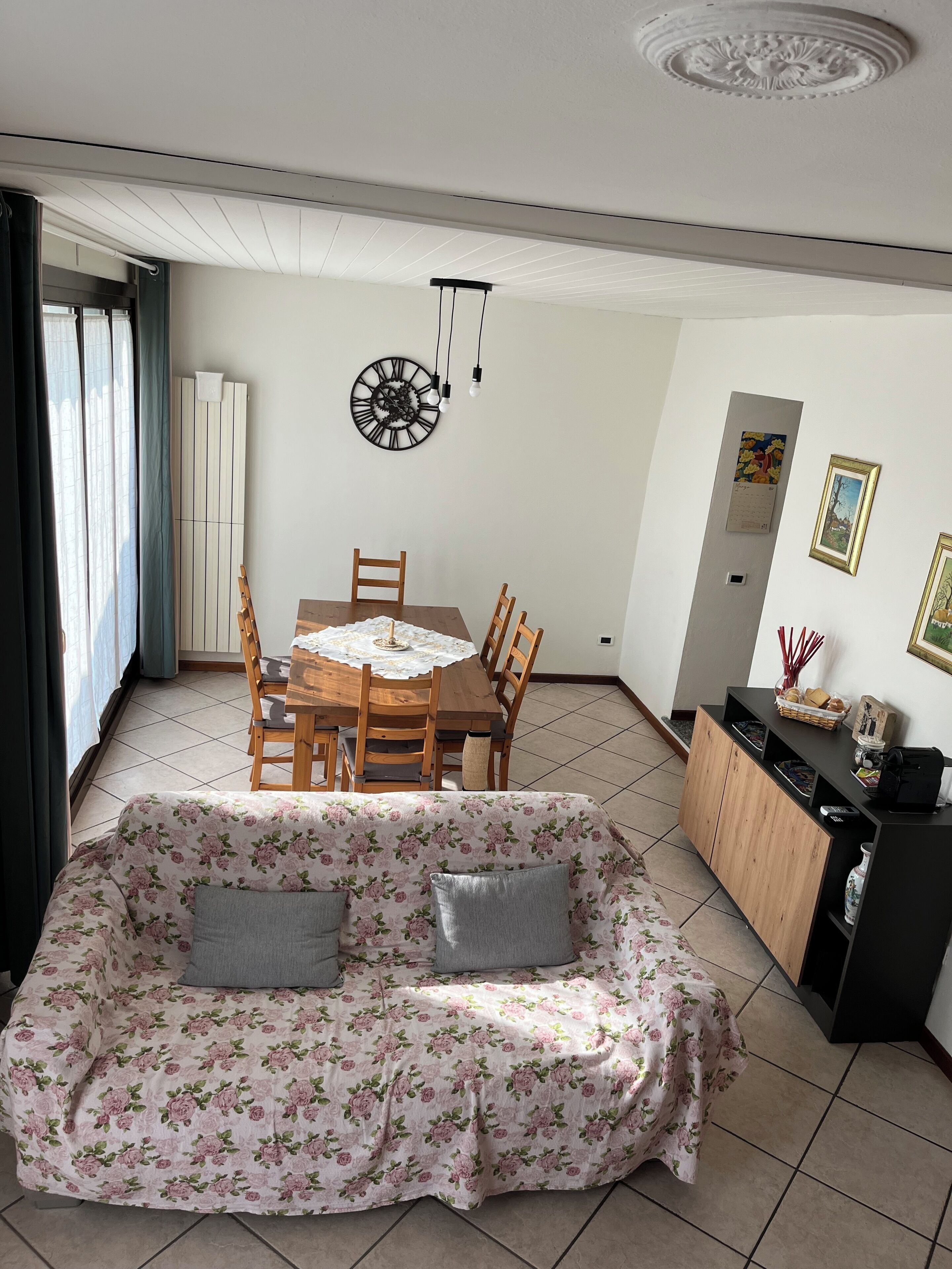 Una sala de estar con un sofá floral, una mesa de comedor con sillas, un armario negro y un reloj de pared.