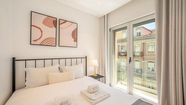 Hotelkamer met een bed, twee kunstwerken aan de muur, een nachtkastje met een lamp en uitzicht op gebouwen door een glazen deur.