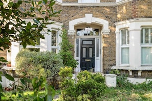 A Victorian-style building with a black front door, a sign above the door, and a small garden area with bushes and a tree. - Charming Victorian Flat with Private Garden in Shepherd's Bush (Greater London)