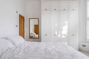 Ein Schlafzimmer mit Bett, weißen Wänden, einer Holztür, einem Kleiderschrank und einem Spiegel.