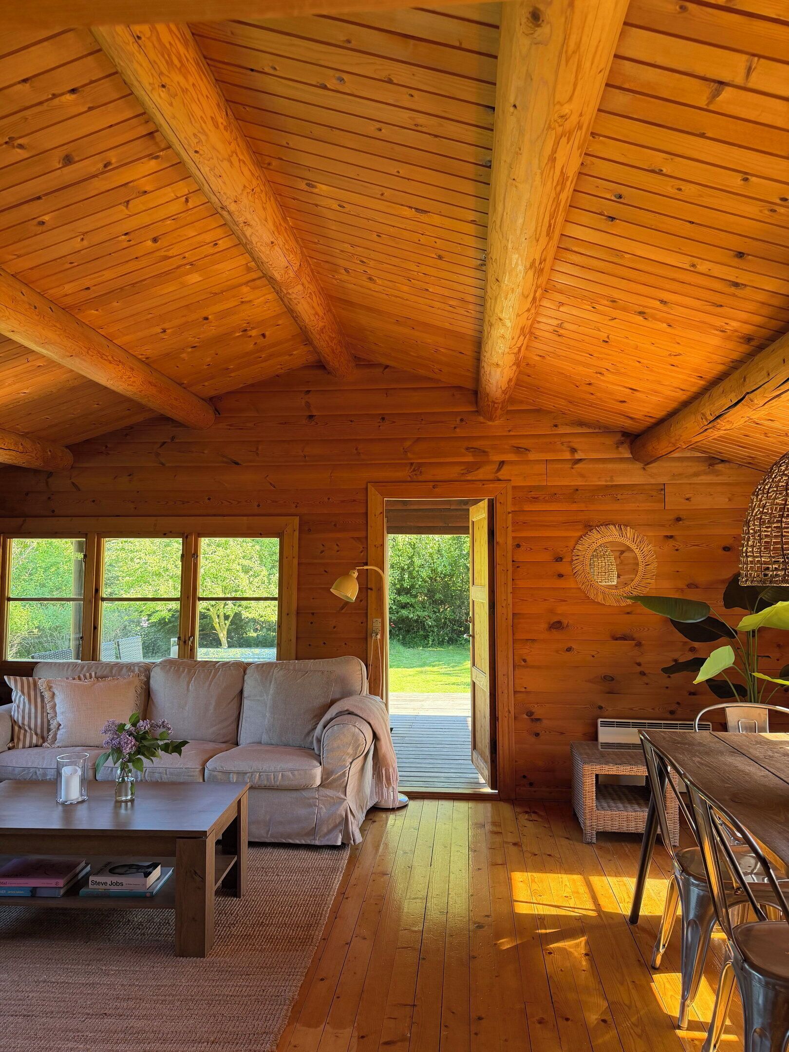 A cozy cabin interior with a wooden ceiling, a sofa, a dining table, and a view of greenery outside.