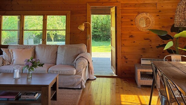 A cozy cabin interior with a wooden ceiling, a sofa, a dining table, and a view of greenery outside. - Charming log cabin in Tisvilde (Tisvilde)