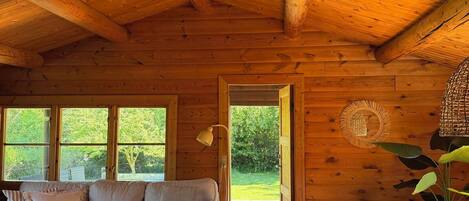 A cozy cabin interior with a wooden ceiling, a sofa, a dining table, and a view of greenery outside.