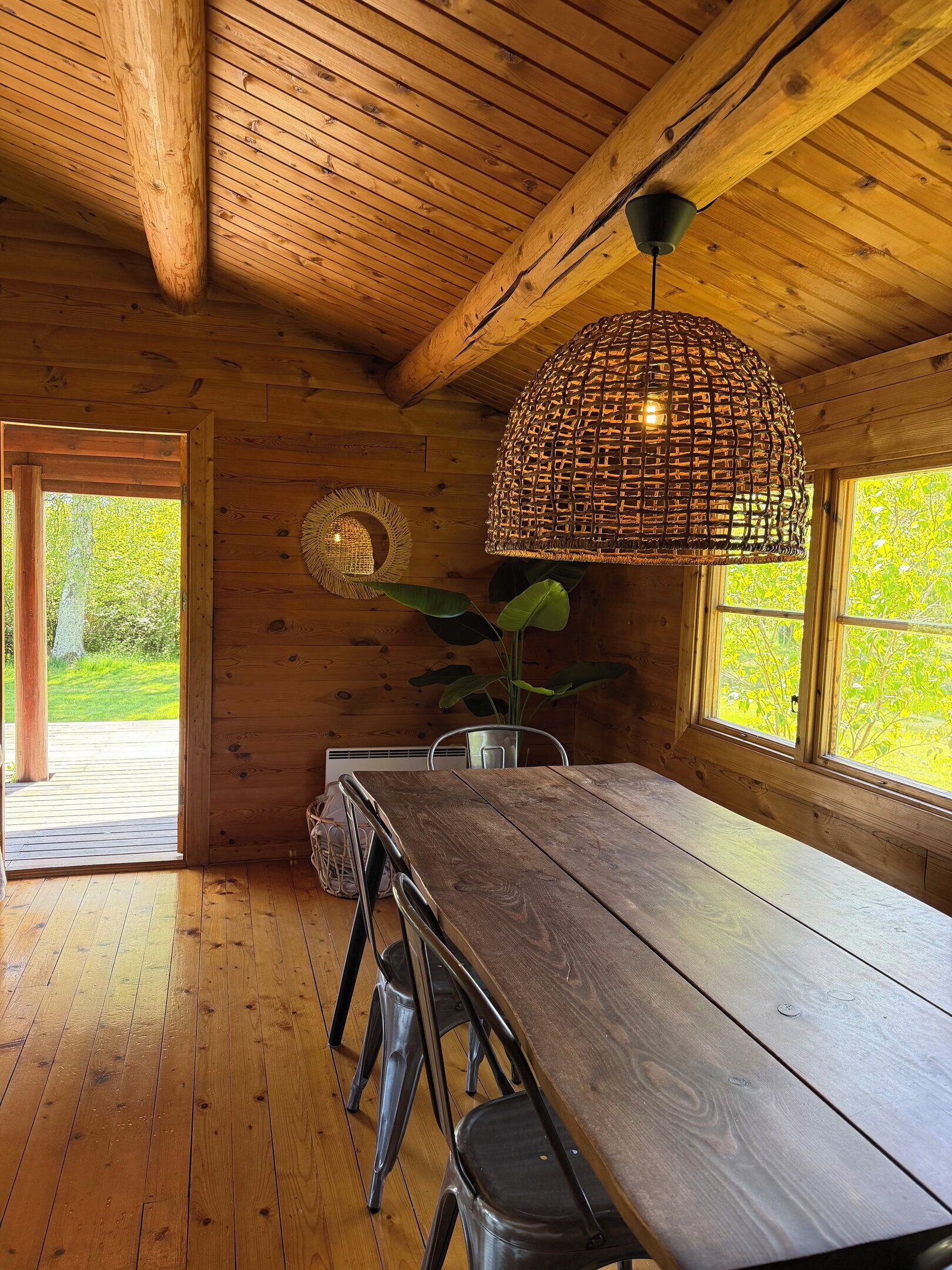 A wooden interior with a dining table, chairs, a hanging light fixture, and a window with a view of greenery.