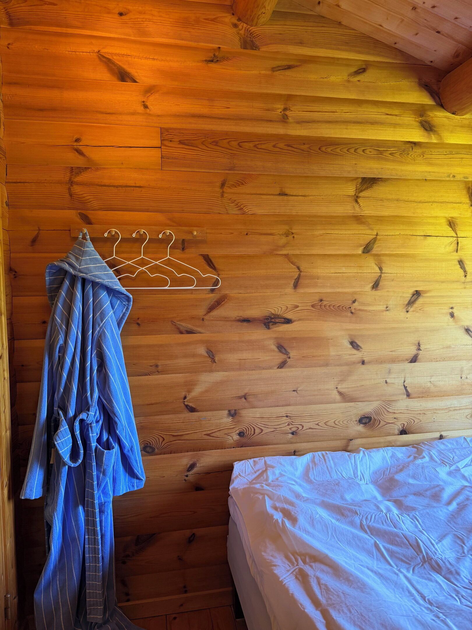 A wooden cabin interior with a bed, a hanging clothes rack, and a wooden ceiling.