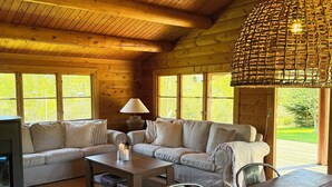 A cozy cabin interior with a wooden ceiling, a woven light fixture, a sofa set, a dining table, and a fireplace. - Charming log cabin in Tisvilde (Tisvilde)